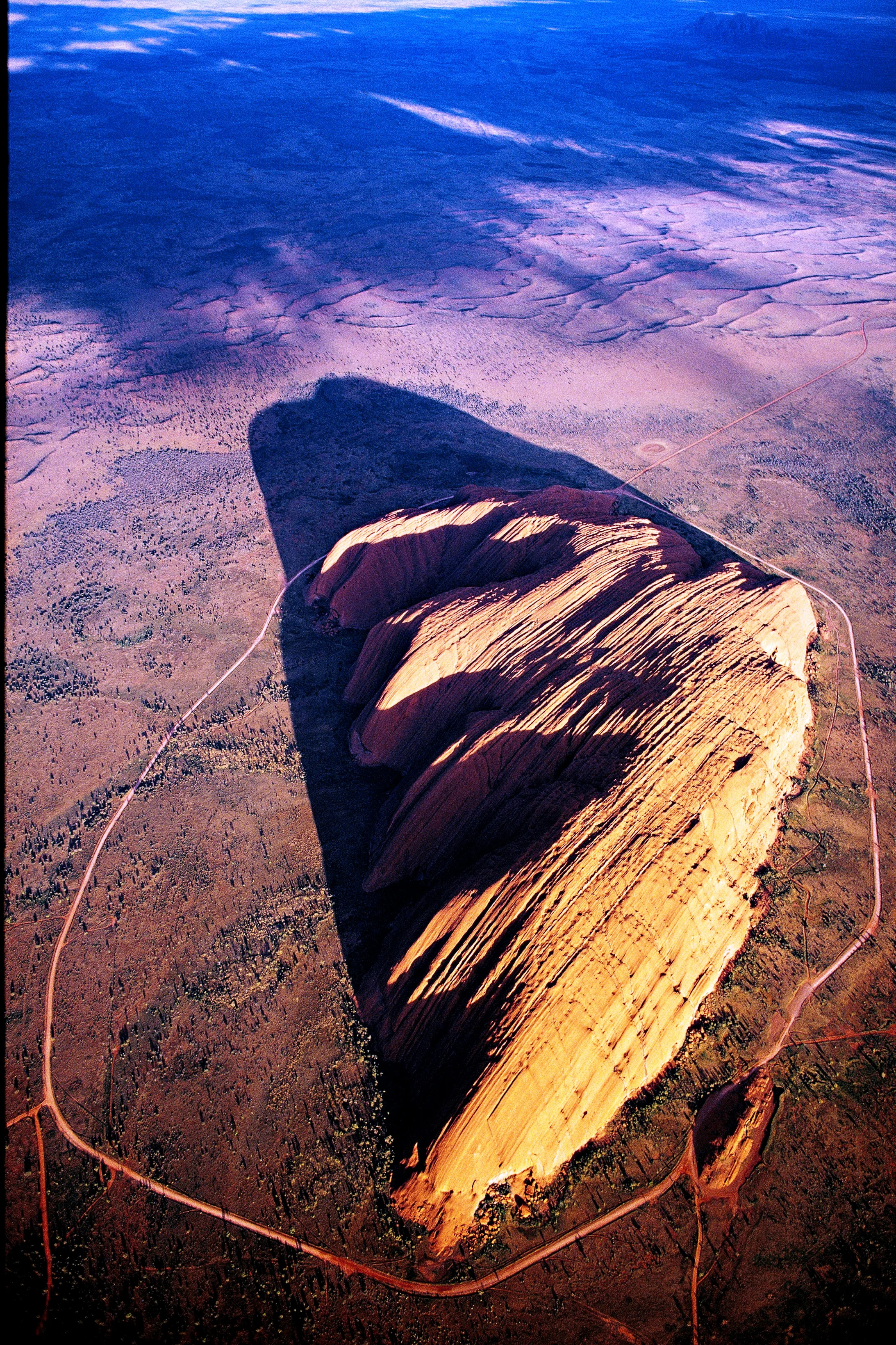 Der Uluru, 348 Meter über die umgebende Wüste emporragend, ist ein Berg der dritten Art: ganz eingesponnen in die „Traumzeit“ der örtlichen Ureinwohner.
Mag ja sein, dass der Fels im roten toten Herzen Australiens schon kraft seiner Natur weltweit einzigartig ist – mitnichten ist er aber ein Monolith, obwohl sogar die Unesco dieses Touristenlatein nachplappert. Und als Inselberg aus geschichtetem Sandstein, den Erosion und Verwitterung aus einer Rumpffläche herauspräparierten, ist er nicht einmal – so ein…